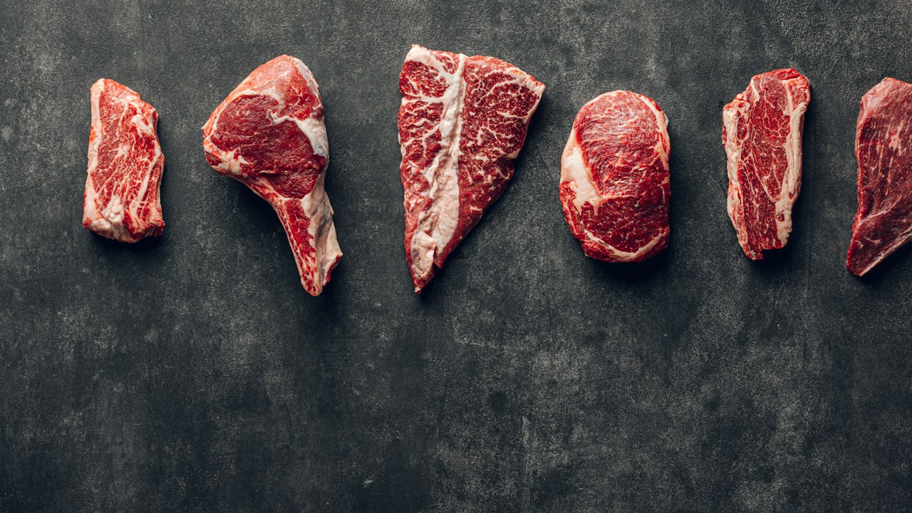 Variedad de cortes de carne vacuna cruda dispuestos sobre una superficie oscura y plana.