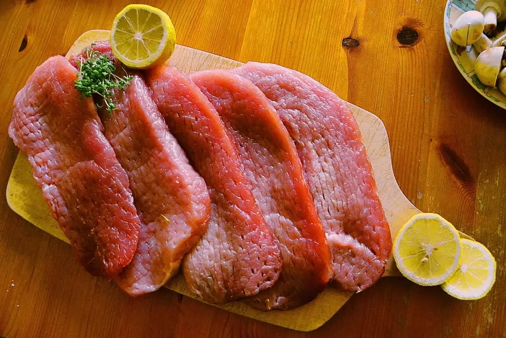 Varios filetes de lengua vacuna cruda dispuestos sobre una tabla de madera con rodajas de limón.