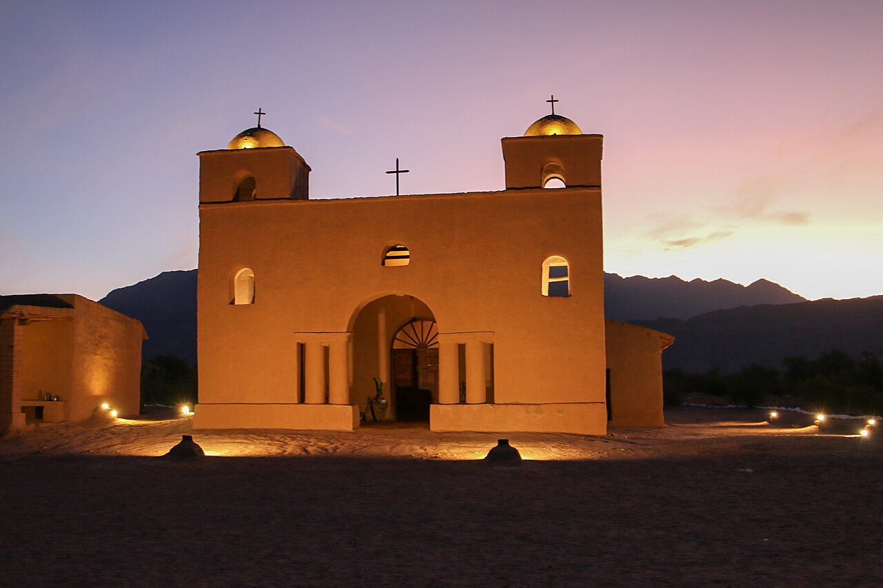 Una iglesia colonial de adobe iluminada al atardecer con montañas en el horizonte lejano.