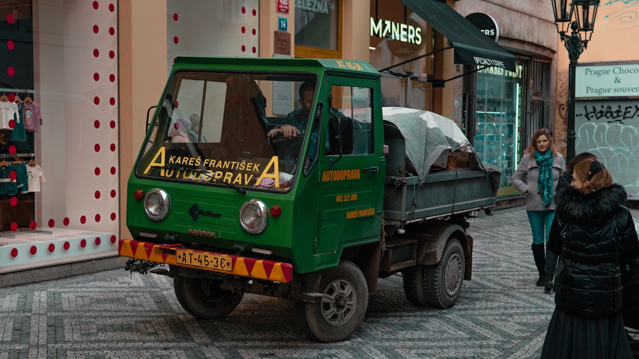Un pequeño camión de carga verde se encuentra estacionado sobre una calle de adoquines europea.