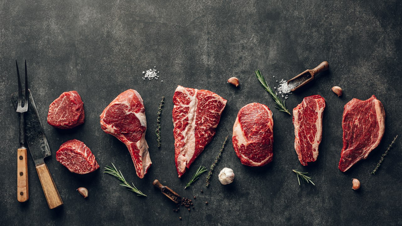 Varias piezas de carne vacuna cruda dispuestas sobre una superficie oscura junto a utensilios de cocina.