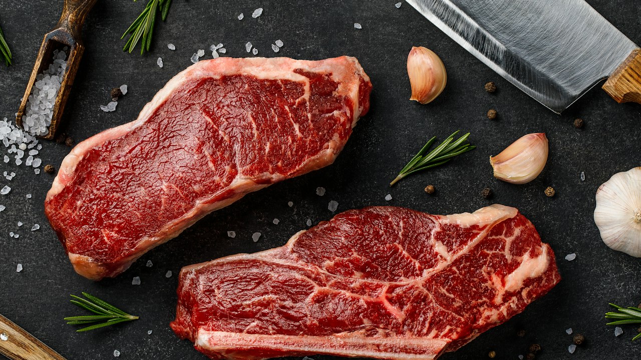 Dos cortes de carne vacuna cruda acompañados de romero, sal, pimienta y un cuchillo de cocina.