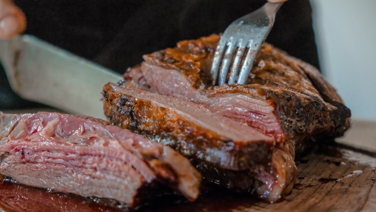 Un cuchillo corta un trozo de carne asada sobre una tabla de madera redonda.
