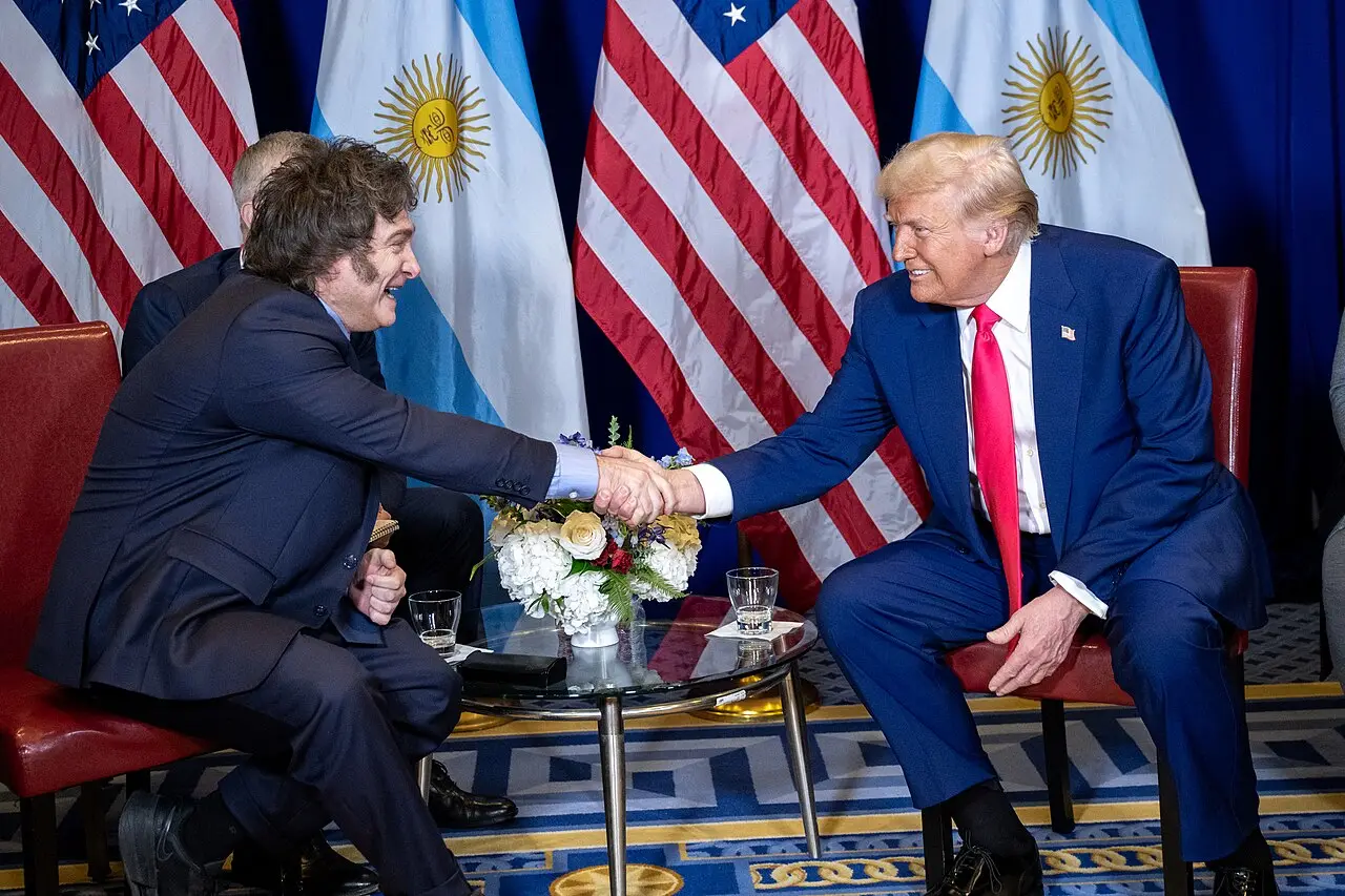 El presidente Javier Milei estrecha la mano de Donald Trump frente a banderas de ambos países.