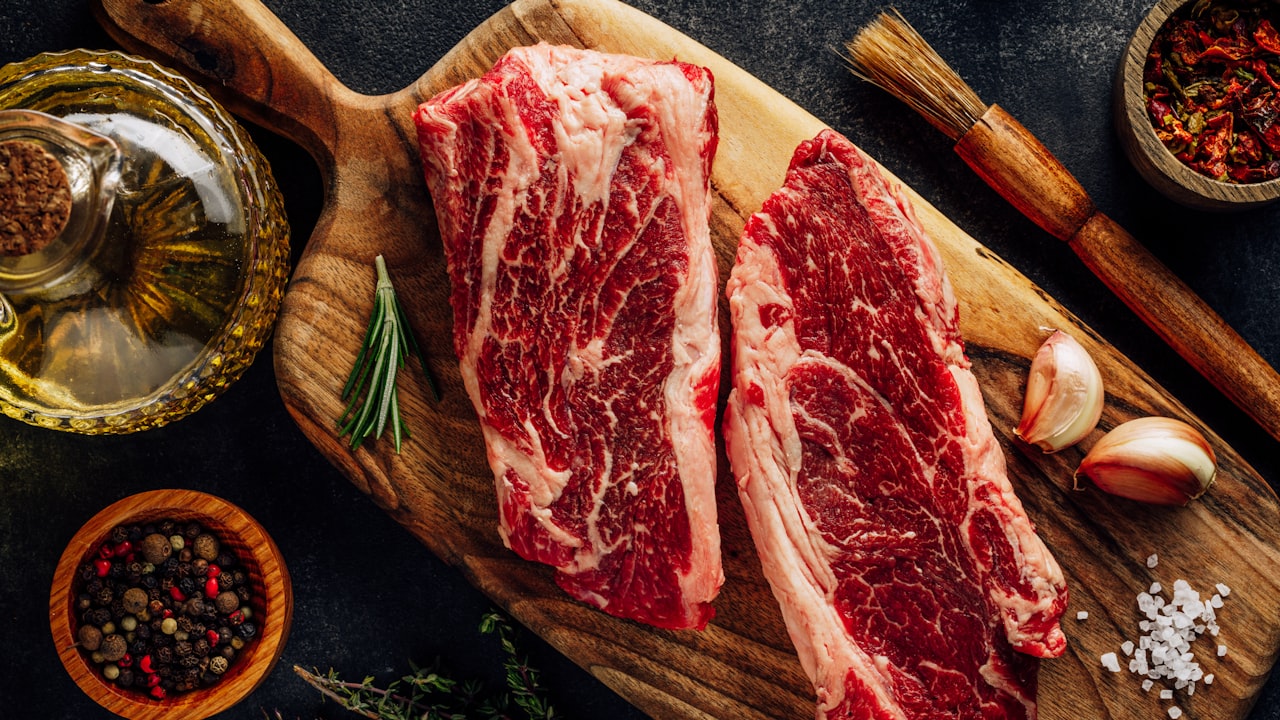 Dos cortes de carne vacuna cruda sobre una tabla de madera con especias y condimentos.