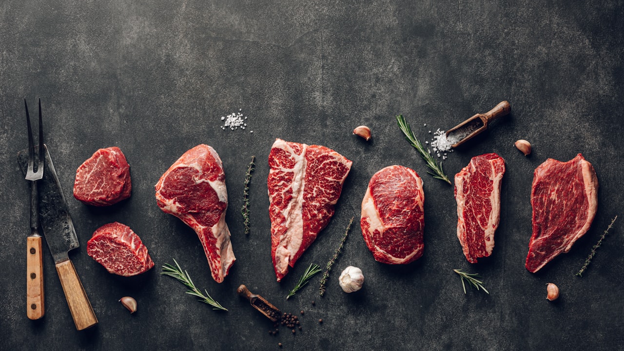 Variedad de cortes de carne cruda dispuestos sobre una superficie oscura con utensilios de cocina.