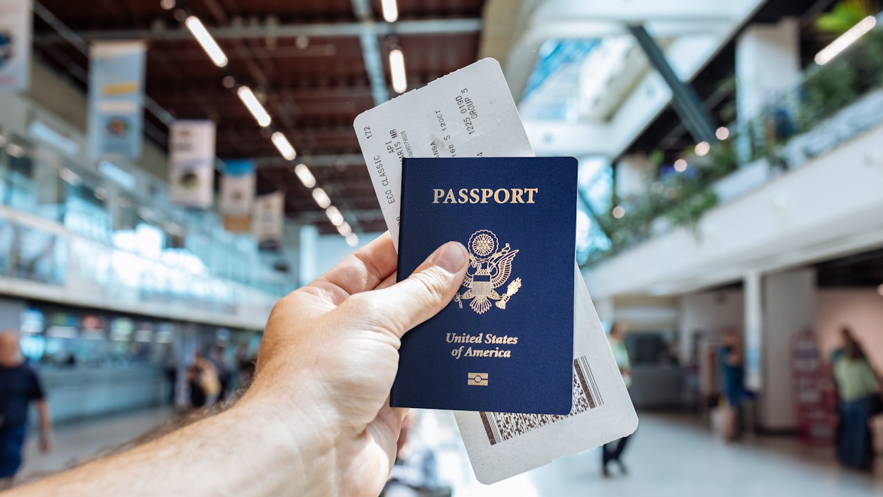 Una mano sostiene un pasaporte estadounidense con tarjetas de embarque en un aeropuerto concurrido.