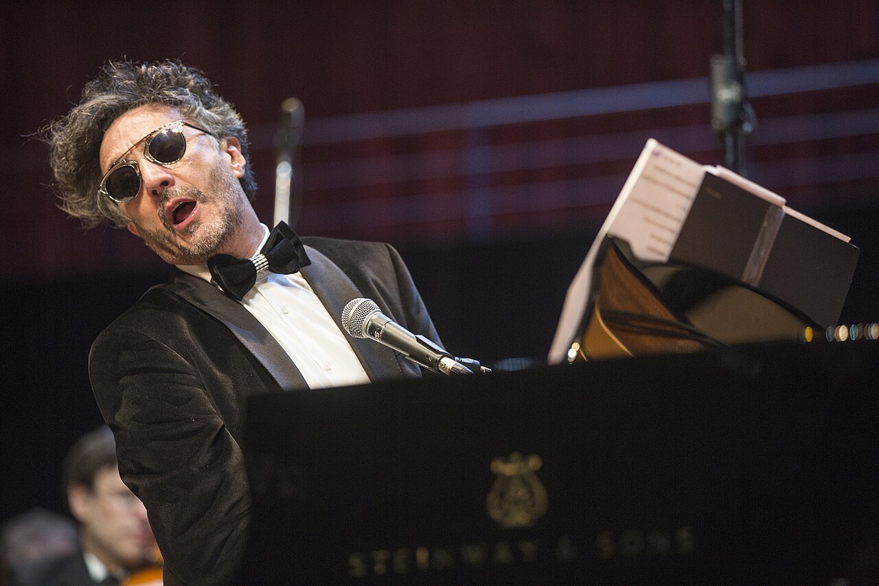 El músico Fito Páez canta frente a un piano de cola en un escenario iluminado.