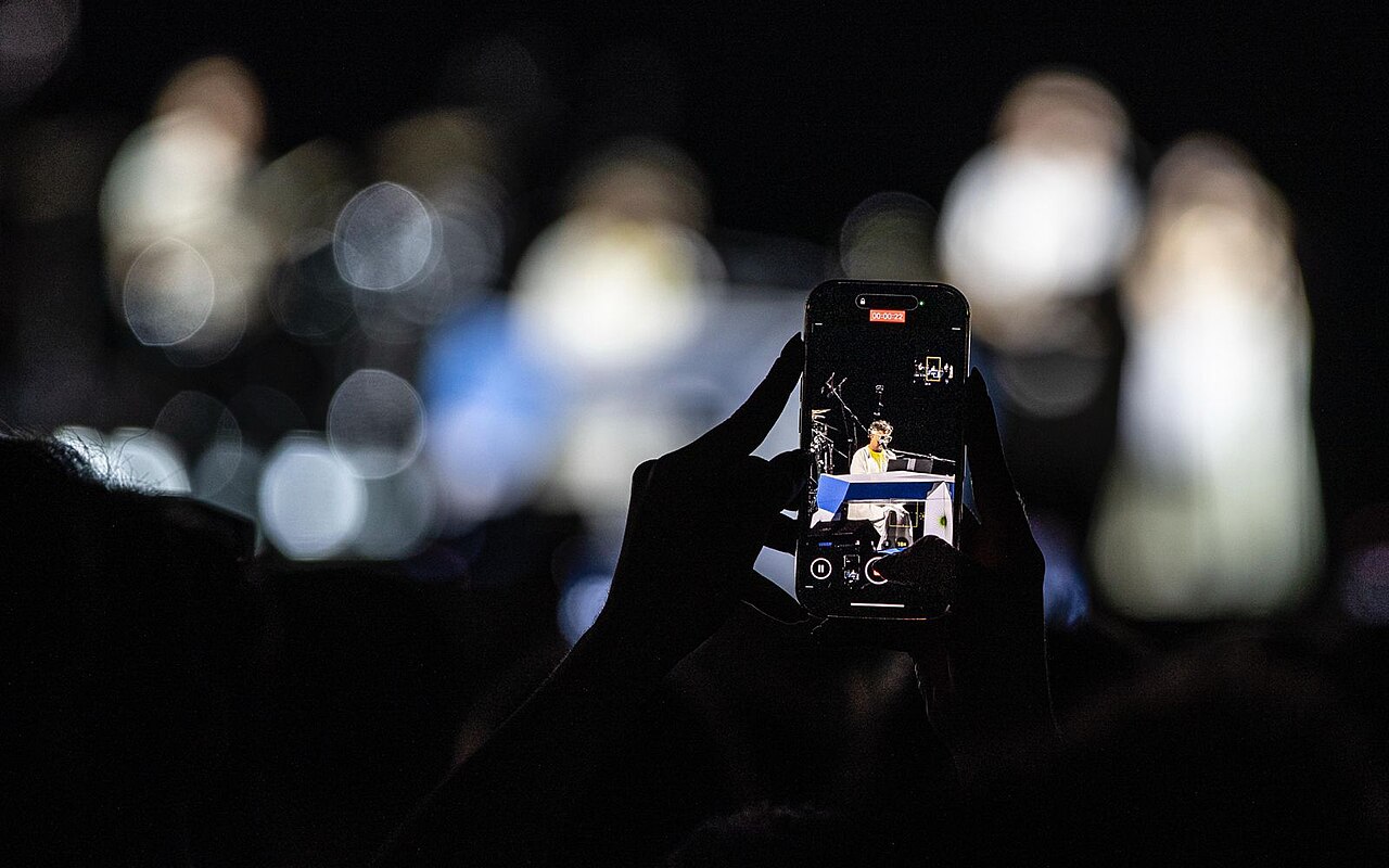 Manos sosteniendo un teléfono celular que graba a un artista sobre un escenario iluminado.