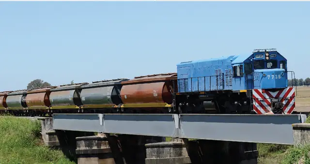 Un tren de carga azul circula sobre un puente ferroviario en una zona rural despejada.