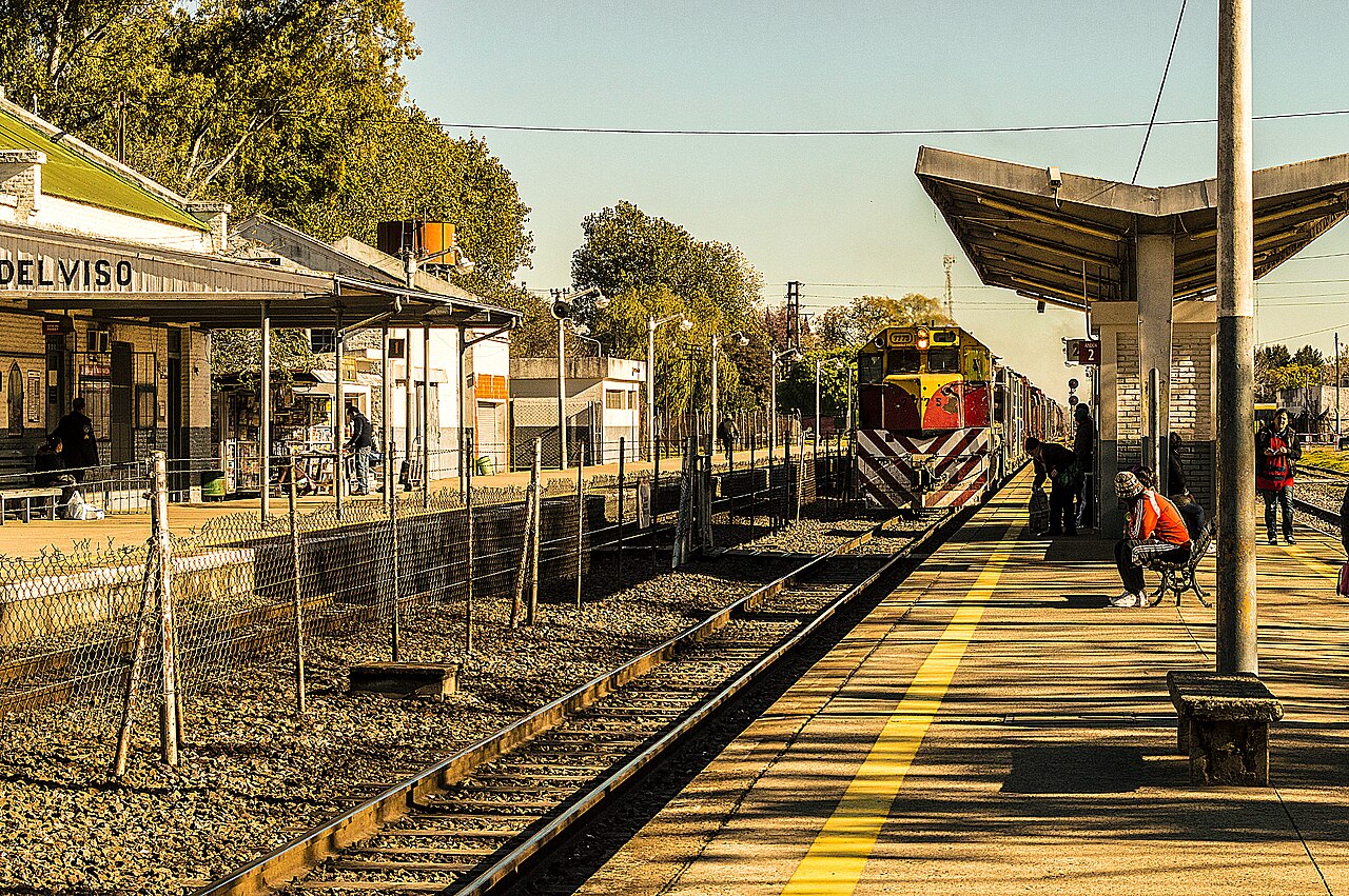 Vista de una estación de tren con pasajeros esperando en el andén bajo el sol.