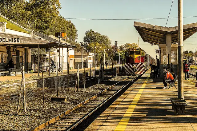 Vista de una estación de tren con pasajeros esperando en el andén bajo el sol.