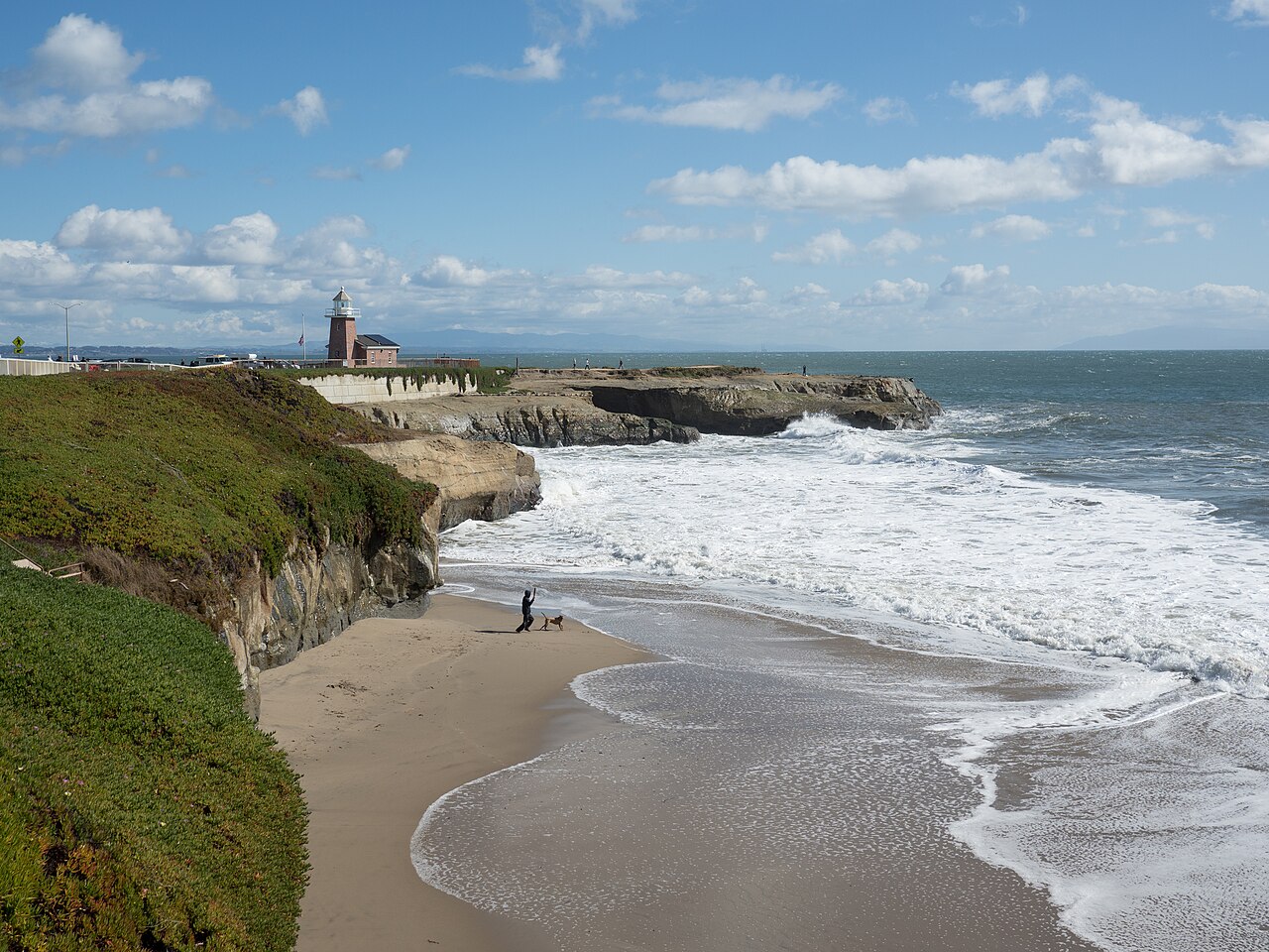 Vista panorámica de una costa con acantilados, un faro y una persona caminando por la playa.