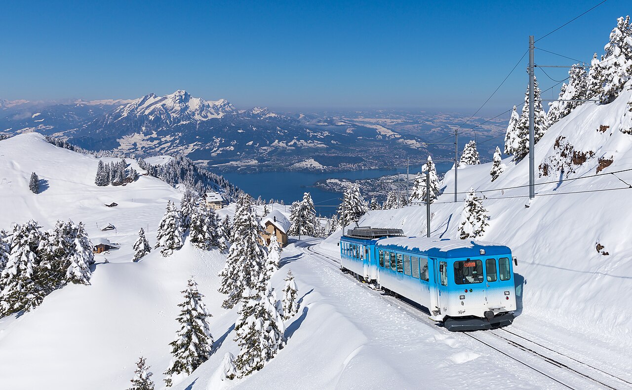 Un tren de montaña recorre un paisaje nevado con montañas y un lago al fondo.