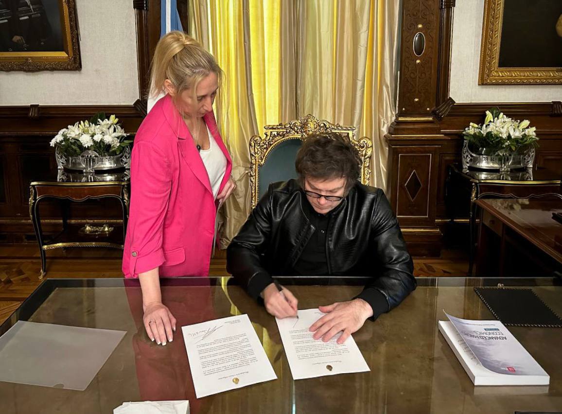 Karina Milei observa a Javier Milei mientras firma documentos oficiales en un escritorio de oficina.