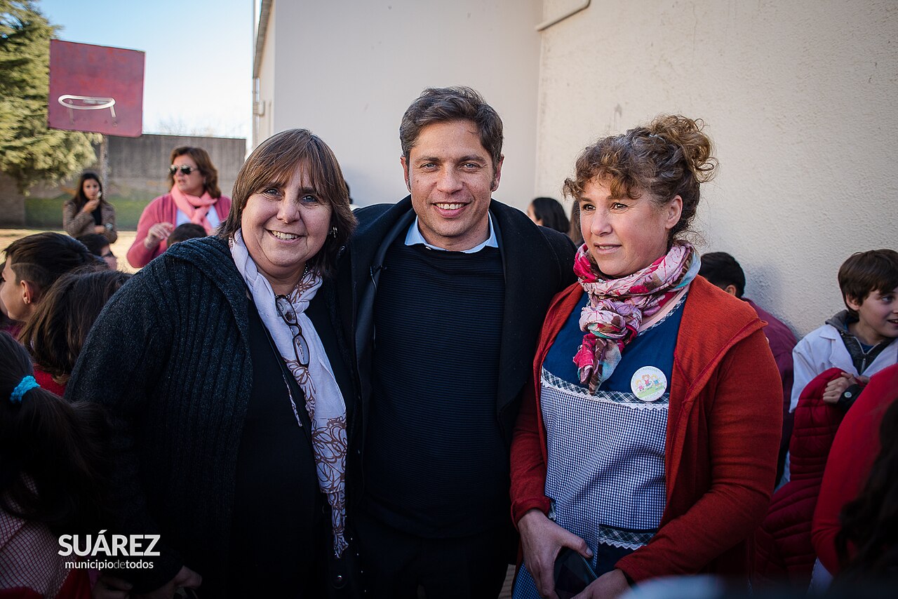 El gobernador Axel Kicillof sonríe flanqueado por dos mujeres en una visita institucional.