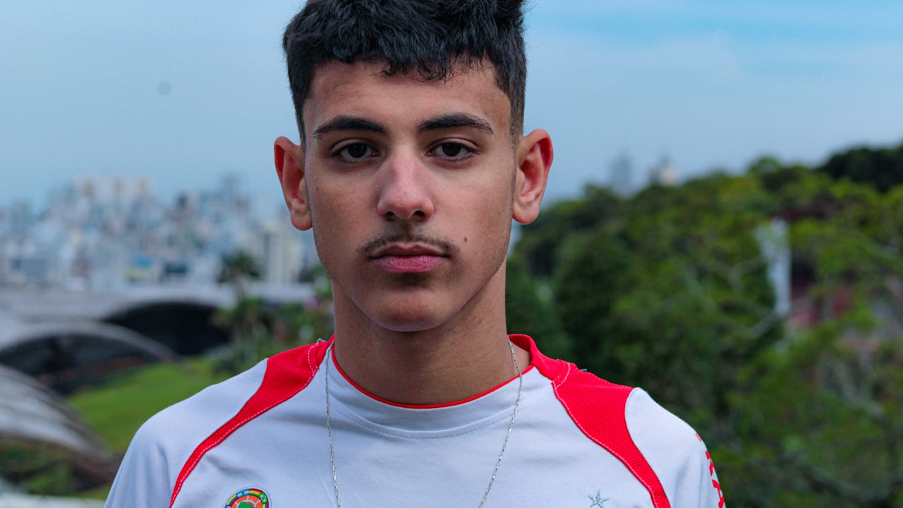 Un joven con bigote y cabello corto posa al aire libre frente a un paisaje verde.