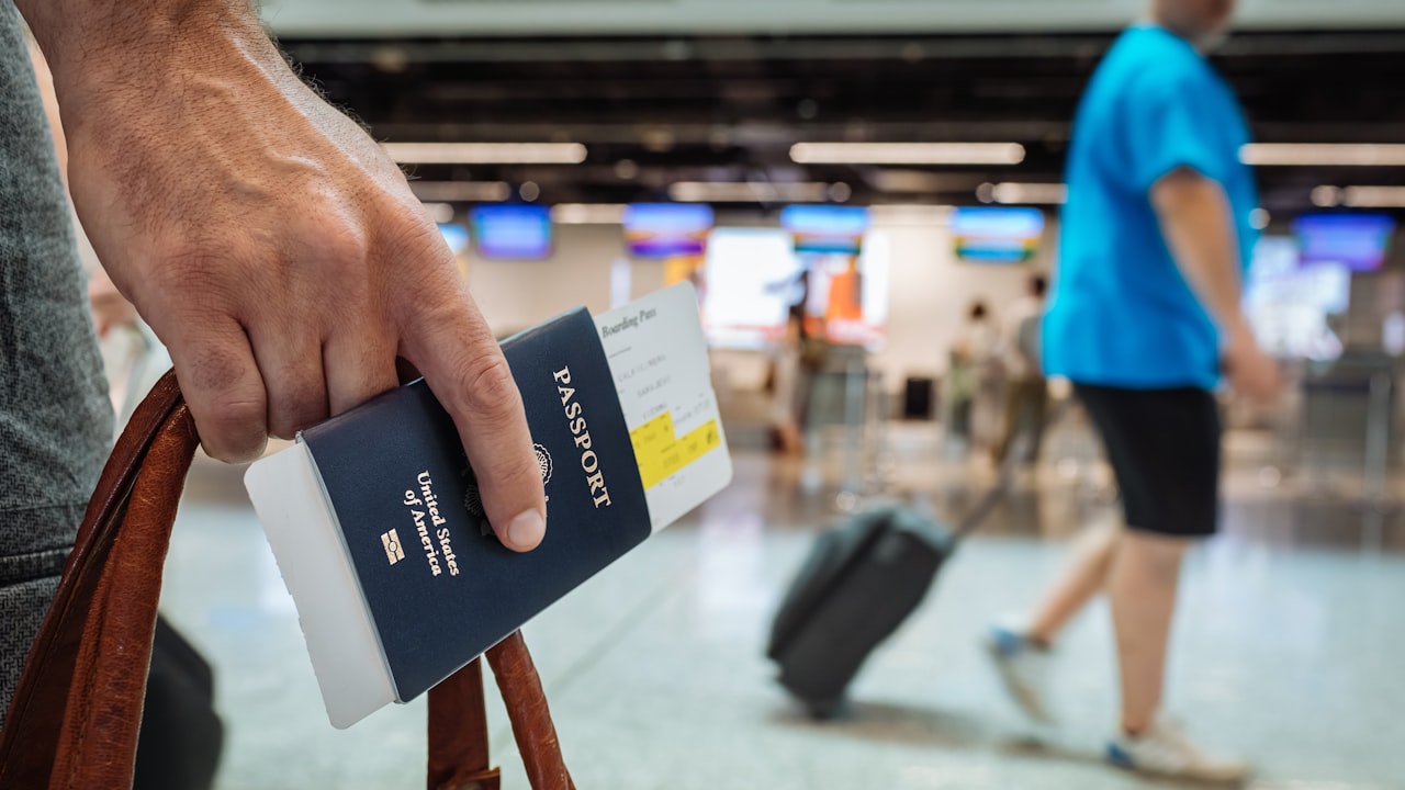 Una mano sostiene un pasaporte estadounidense y un maletín en un aeropuerto con gente caminando.