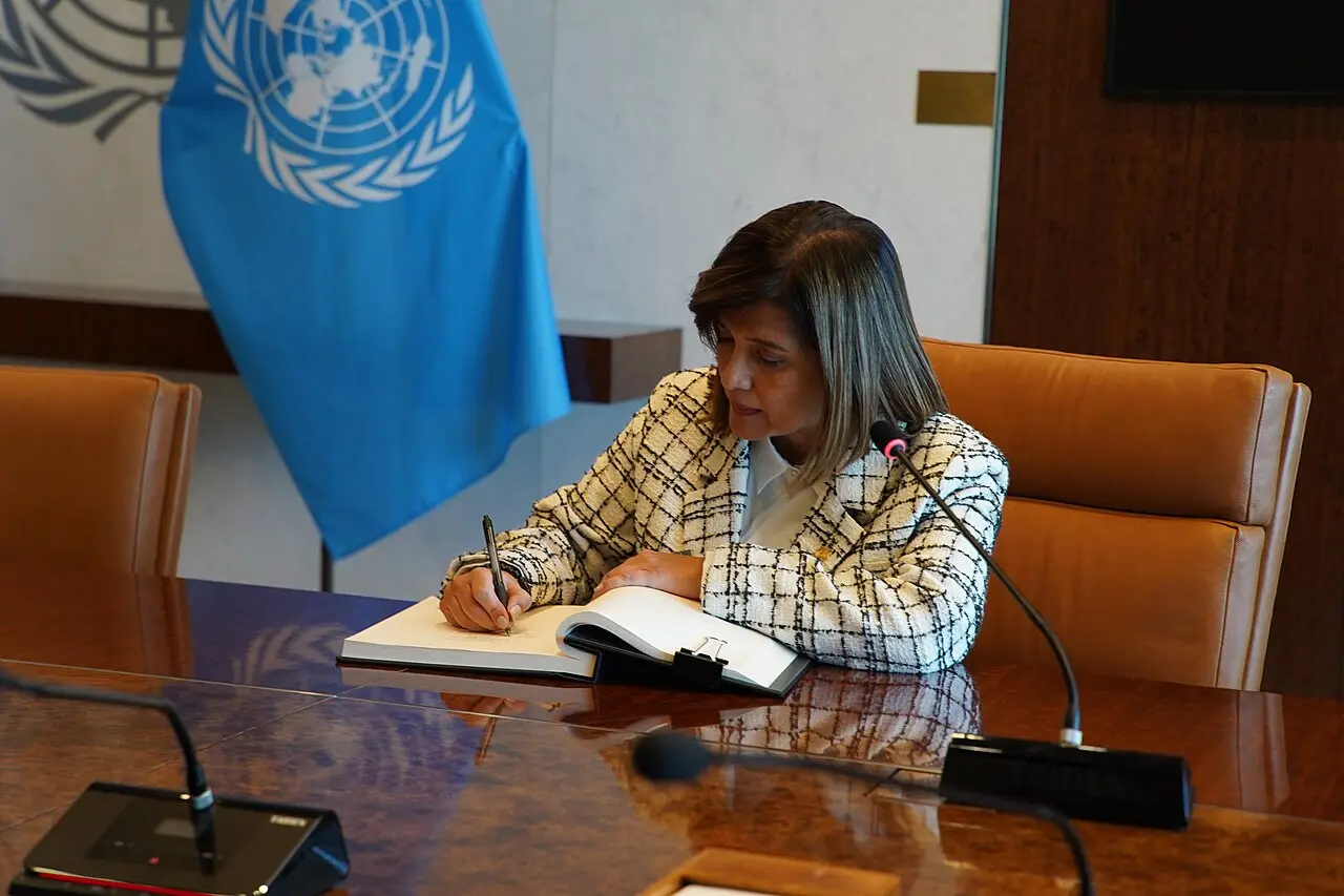 Una mujer firma un documento oficial frente a la bandera de la Organización de Naciones Unidas.