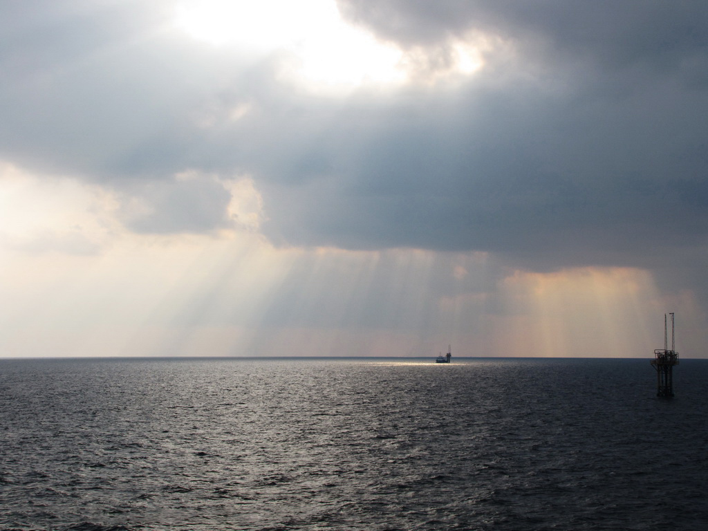 Rayos de sol atraviesan nubes sobre el mar con una plataforma petrolera a lo lejos.